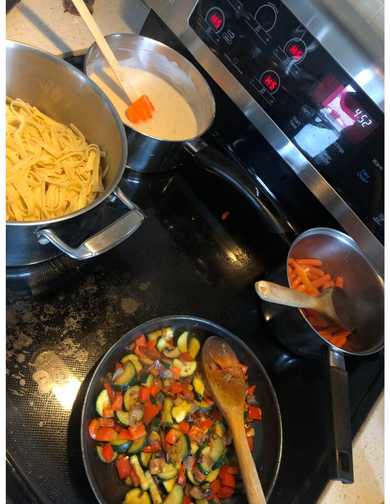 Stovetop with cooked Fettuccine in the bottom left corner and homemade Alfredo sauce in the top left corner. There are also garlic butter carrots in the top right corner and cooked veggies in the bottom right corner.