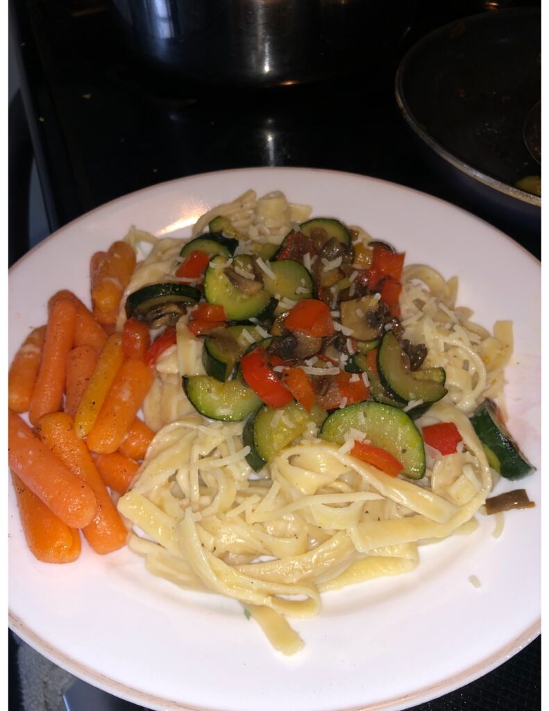 Plate of Fettuccine Alfredo pasta with a side of garlic butter carrots. The pasta is topped with cooked zucchini, mushrooms, and bell peppers.