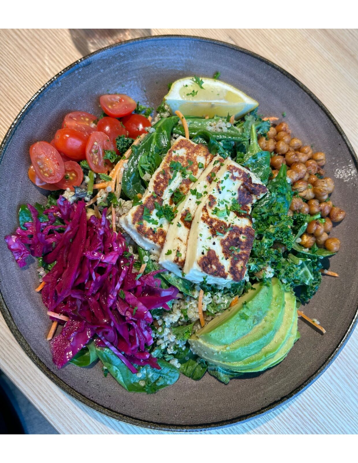 Picture of a quinoa bowl with purple cabbage, avocado, halloumi cheese, kale, chickpeas, cherry tomatoes, and kale. There are also bits of carrots sprinkled across the bowl, with a lime wedge at the top of the bowl.
