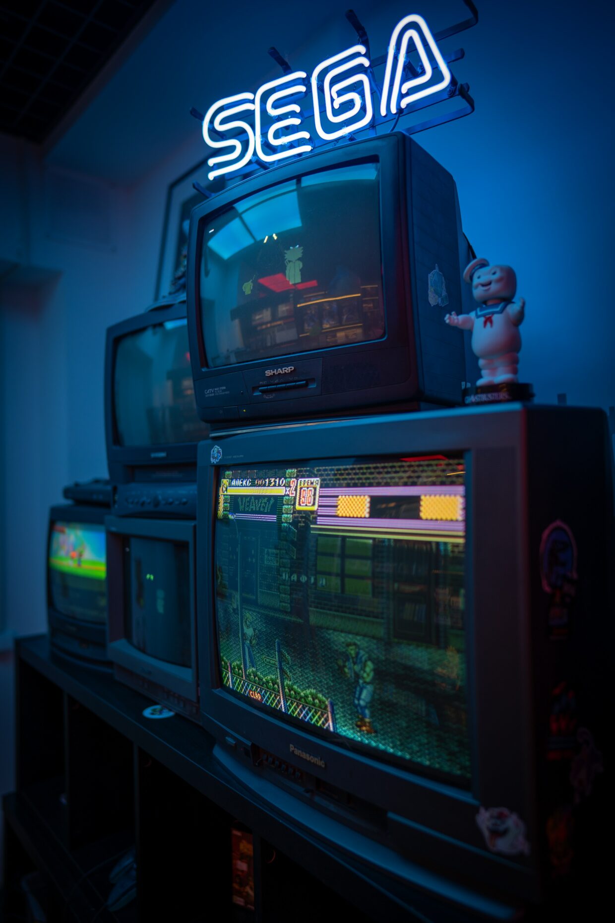 Picture of multiple box-style TV's stacked on one another. There is the SEGA sign above in blue neon lights. The TV screens depict either faded and static imagery or are completely off. Photo by Ivan Rudoy on Unsplash