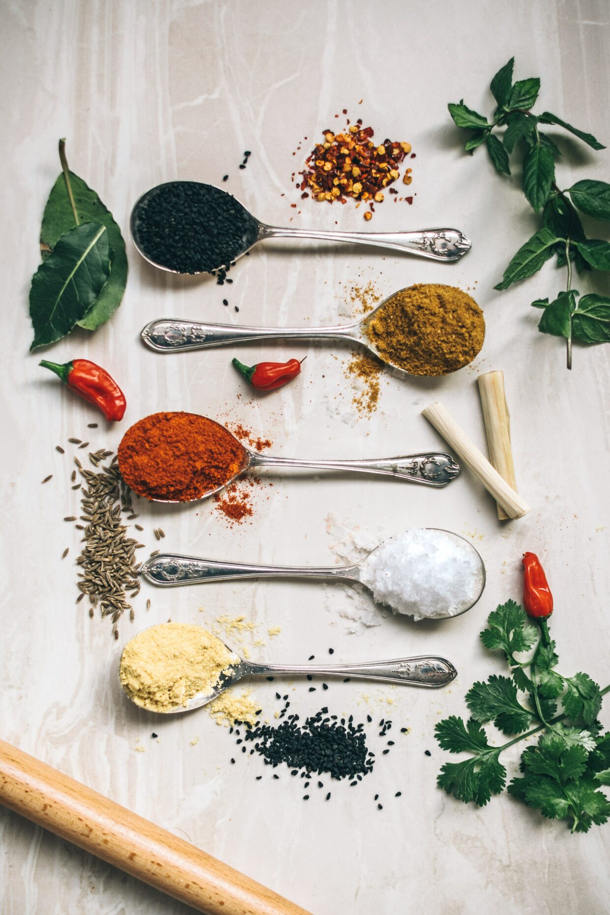 Picture of 5 different spoons filled with spices. Starting from the top going down, there is black sesame seeds, cumin powder, paprika, salt, and garlic powder. These are simply assumptions. There are also various garnishes surrounding the spoons such as cilantro and mint.