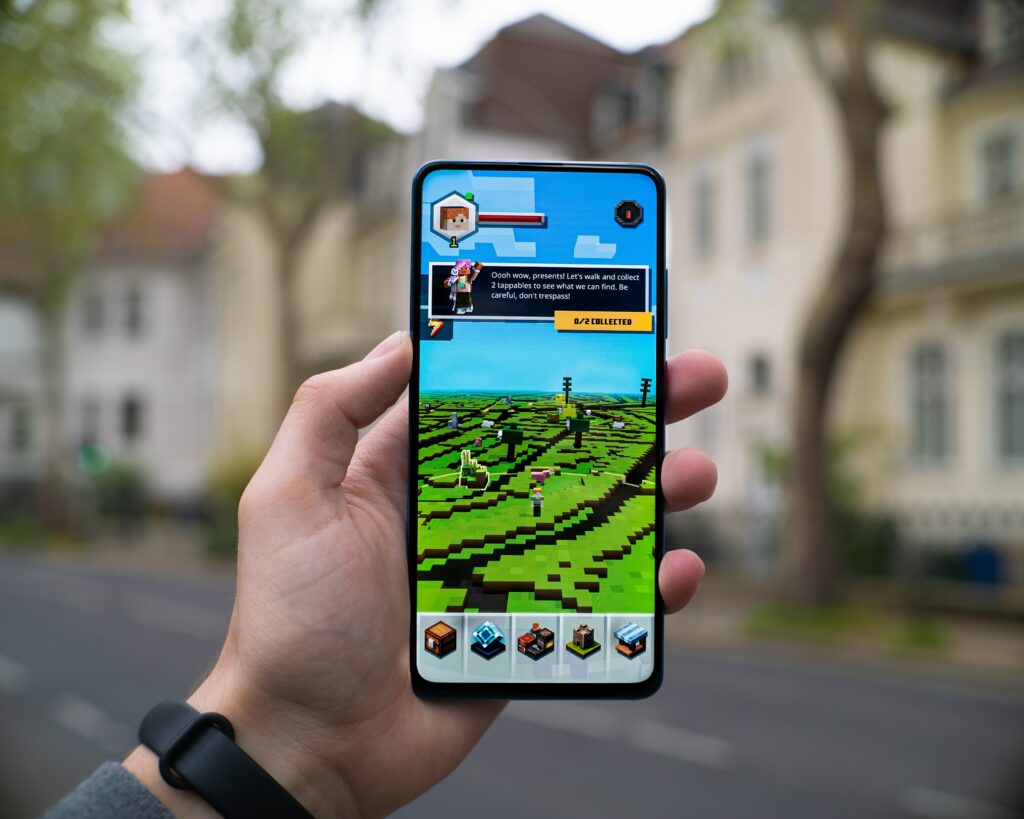 Image of a person holding a phone. On that phone, on the screen, is the game Minecraft. If you look closer, there is a little person with trees going into the distance. 